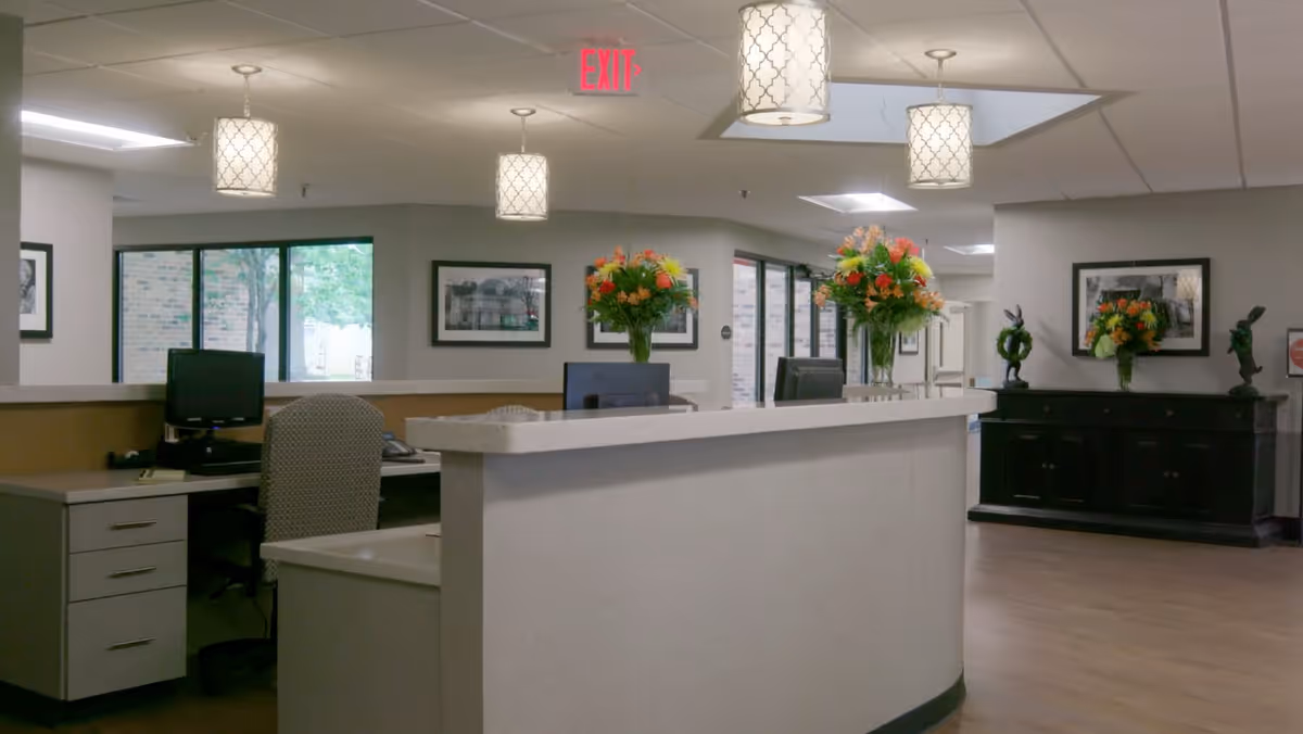 Reception area inside Elk River Health and Nursing Center of Winchester with a curved white desk, computer monitors, office chairs, and decorative flower arrangements. The space has hanging patterned light fixtures, framed black and white photos on the walls, and large windows showing an outside view.
