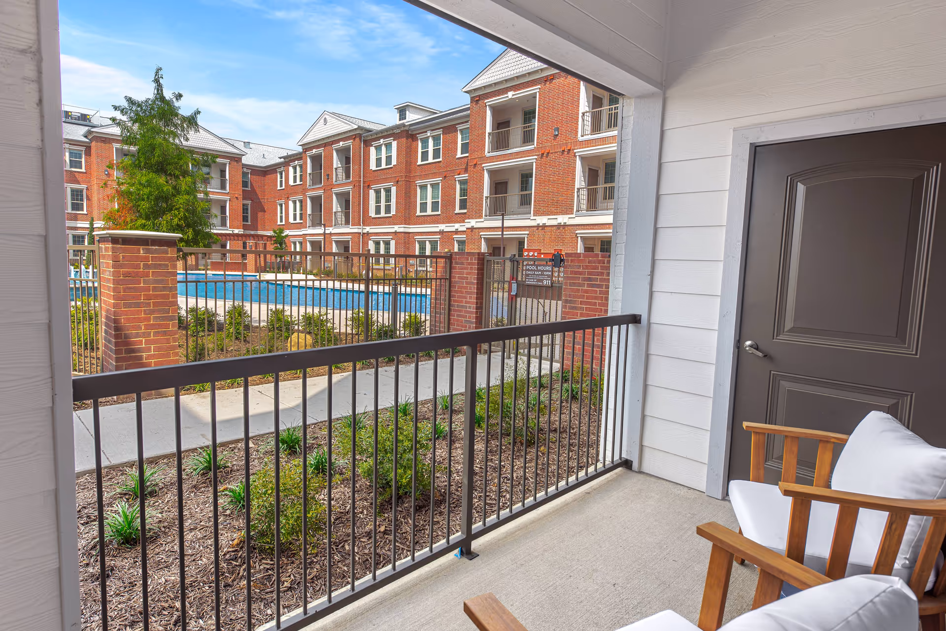 Covered patio with wooden chairs overlooking a fenced pool and red-brick apartment buildings.