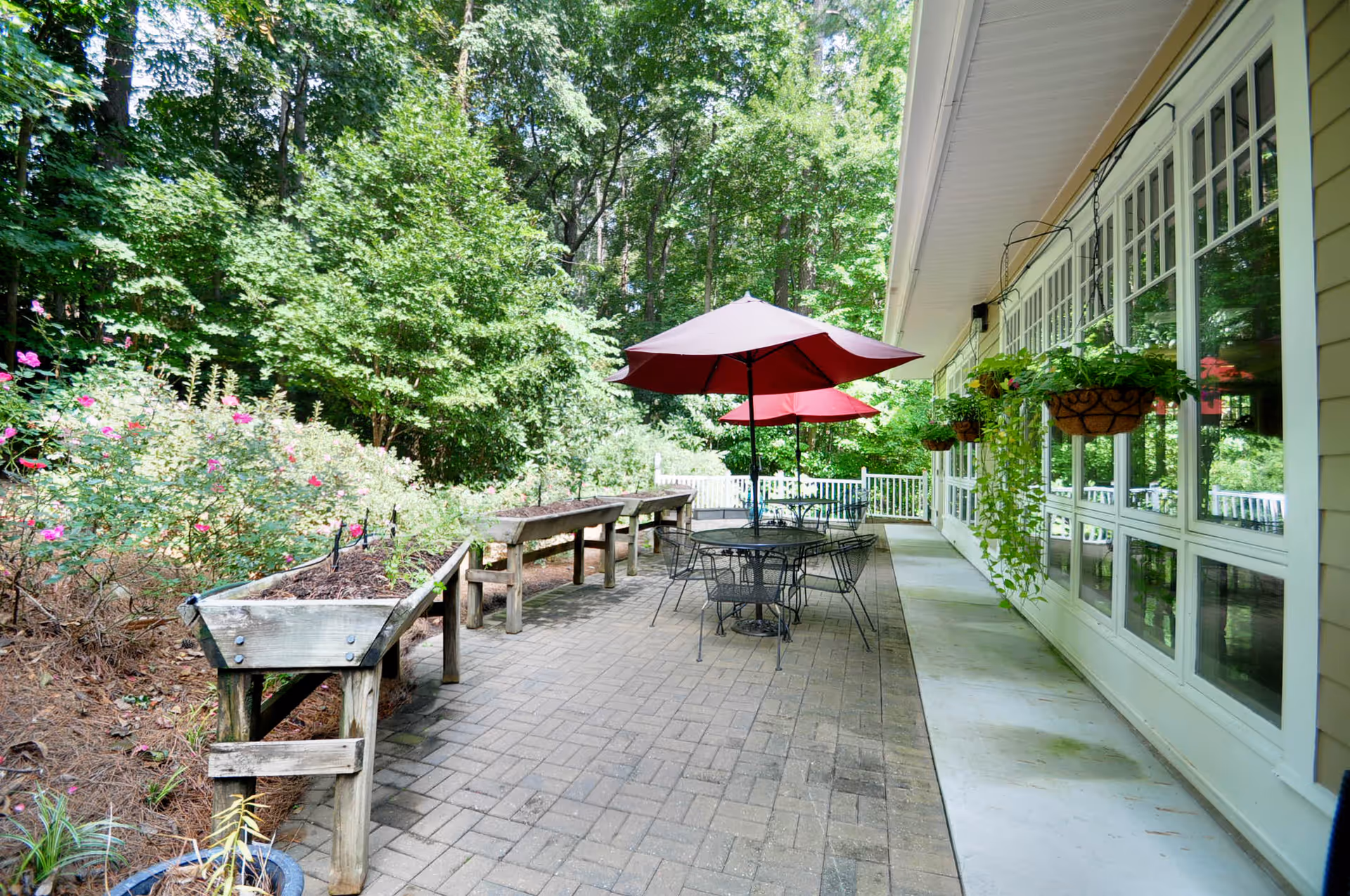Outdoor patio area with brick flooring, featuring black metal tables and chairs under red umbrellas. Raised wooden garden beds with plants and flowers are on the left side, and hanging flower baskets are attached to the exterior wall of a building with large windows on the right. The background shows dense green trees and foliage.
