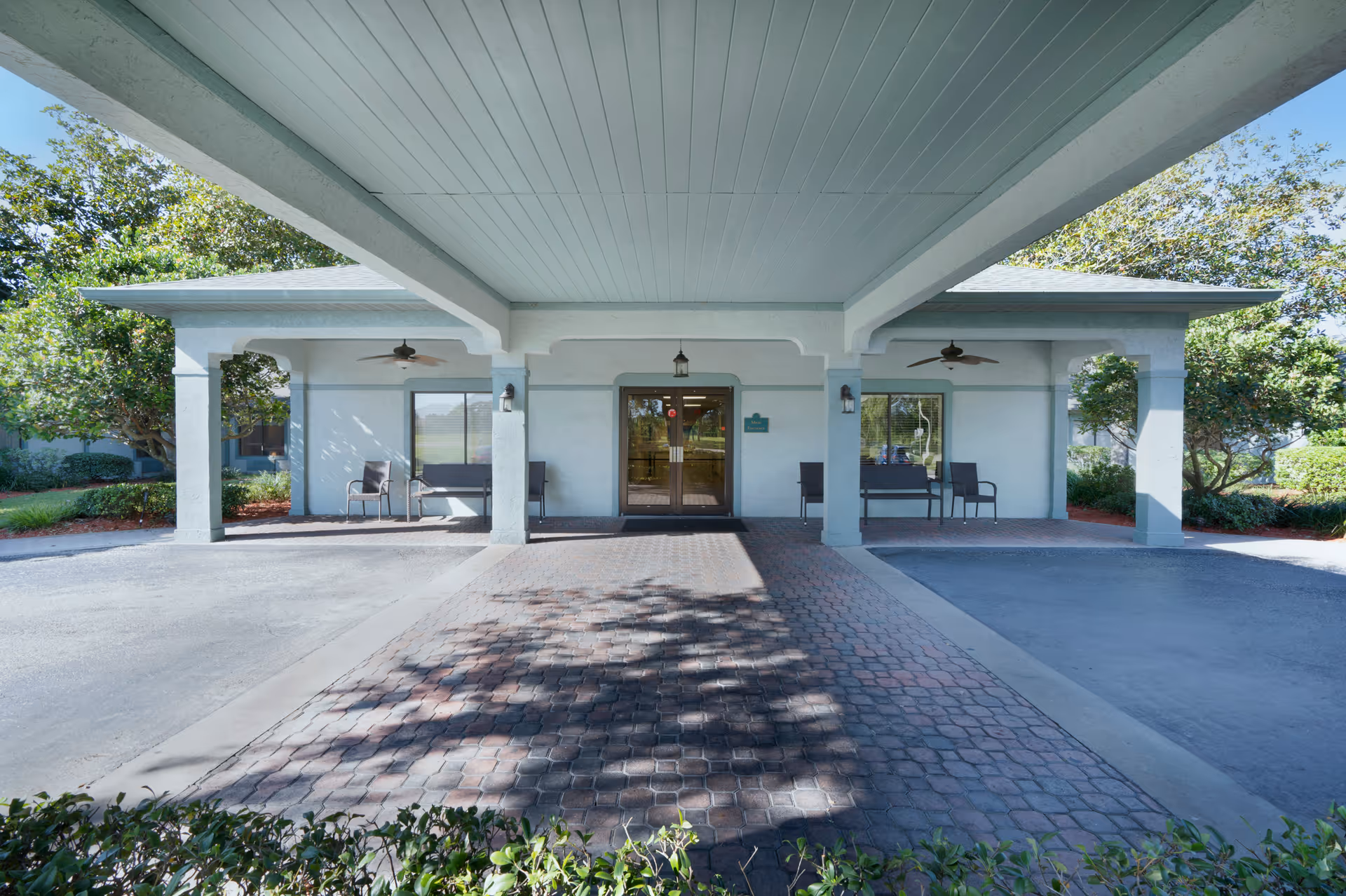 Covered entrance area of Bayonet Point Health Center with a paved driveway leading to double glass doors. The entrance is flanked by seating areas with chairs and benches under a roof supported by columns. Trees and greenery surround the building.