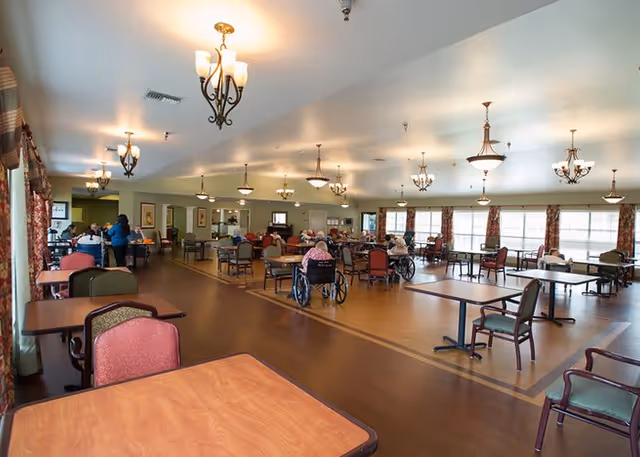 Spacious dining room in a senior living facility with multiple tables and chairs. Several elderly residents, some in wheelchairs, are seated and engaged in activities or conversations. The room is well-lit with large windows along one side and multiple ceiling light fixtures. Curtains with a floral pattern adorn the windows.