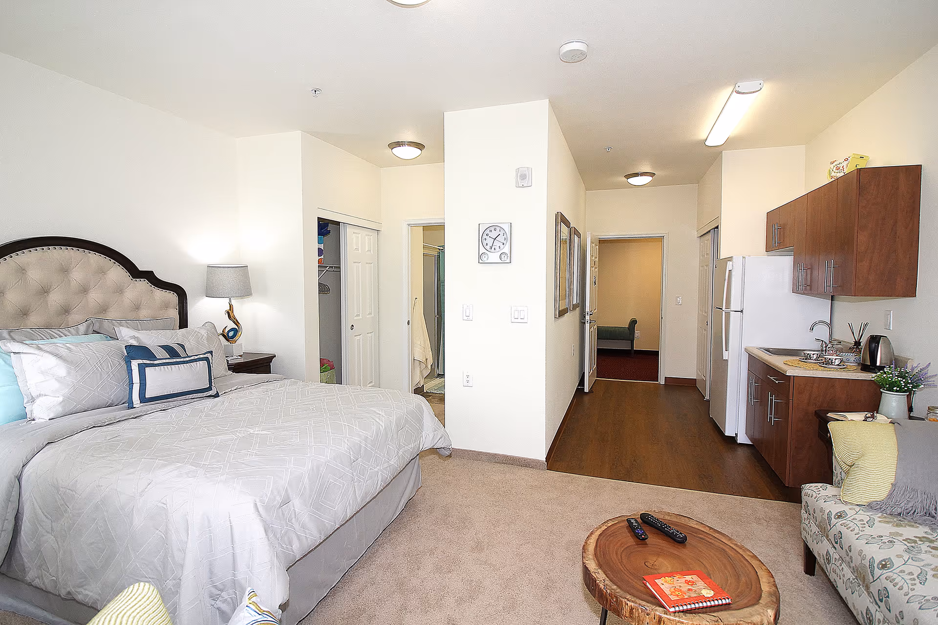 A bright and clean studio apartment featuring a large bed with a tufted headboard, a bedside table with a lamp, a small kitchenette with wooden cabinets, a refrigerator, and a sink. There is a cozy sitting area with a patterned armchair and a wooden coffee table with remote controls and a notebook on it. The room has light-colored walls, carpeted flooring near the bed, and wood flooring near the kitchenette. A hallway leads to a bathroom and closet area.