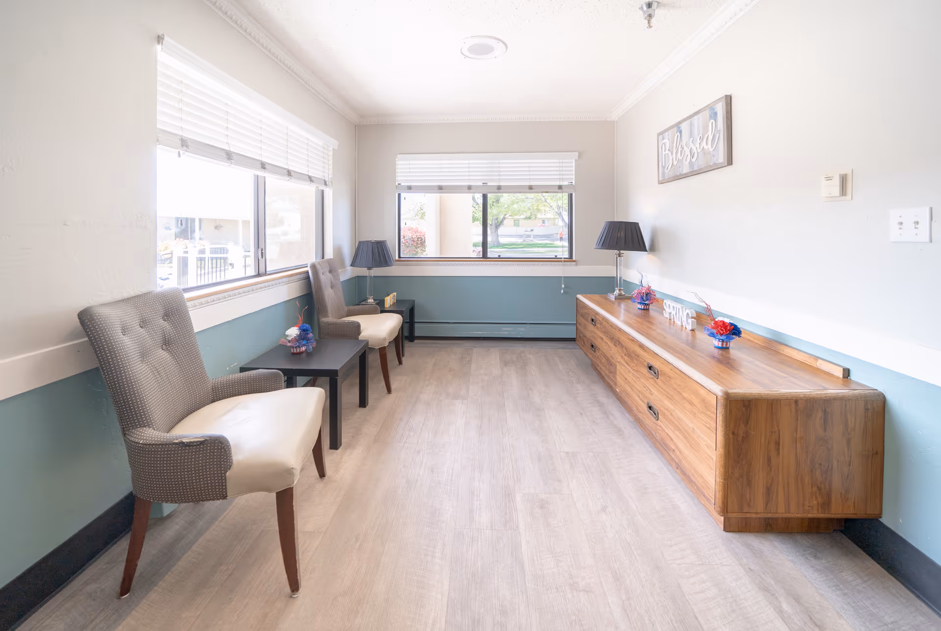 A bright and clean waiting area with two upholstered chairs and a small black table between them. There is a long wooden cabinet on the opposite wall with decorative items including a lamp, flowers, and a sign that says 'SPRING'. Two large windows with white blinds allow natural light to fill the room. A framed sign on the wall reads 'Blessed'.