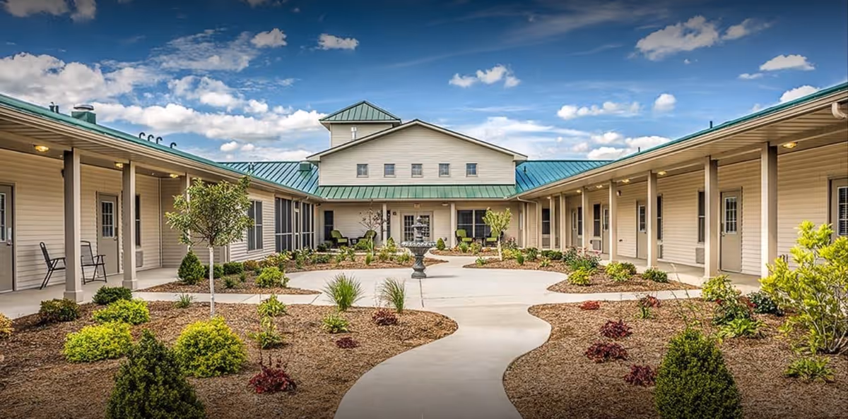 Outdoor courtyard area of Carriage Crossing Senior Living of Arcola featuring a central walkway surrounded by landscaped garden beds with shrubs and small trees, flanked by single-story buildings with beige siding and green metal roofs under a partly cloudy blue sky.