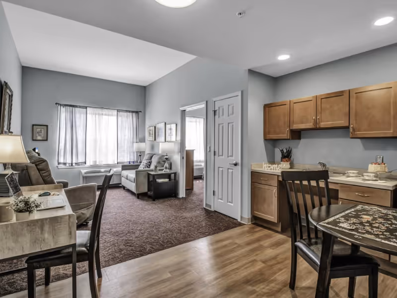 Interior view of a senior living apartment with a small kitchen area featuring wooden cabinets and a round dining table with chairs. The living area has a sofa, armchair, side tables with lamps, and a window with sheer curtains. A doorway leads to a bedroom with a bed visible.