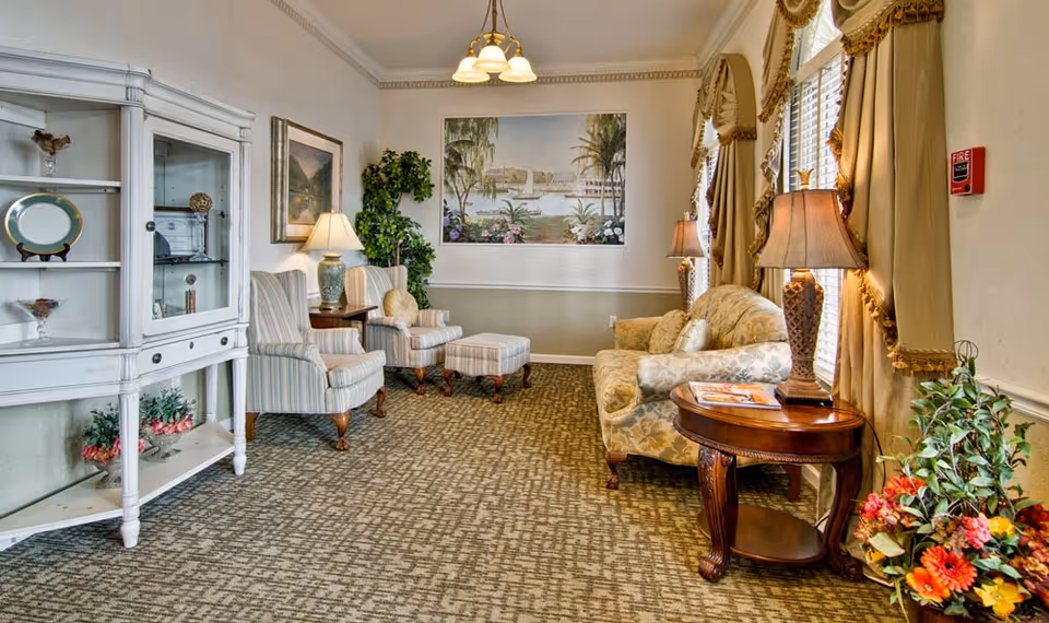 A cozy senior living facility lounge with patterned carpet, a floral sofa, two striped armchairs with a matching ottoman, wooden side tables with lamps, a white display cabinet, framed artwork on the walls, and large windows with beige curtains.