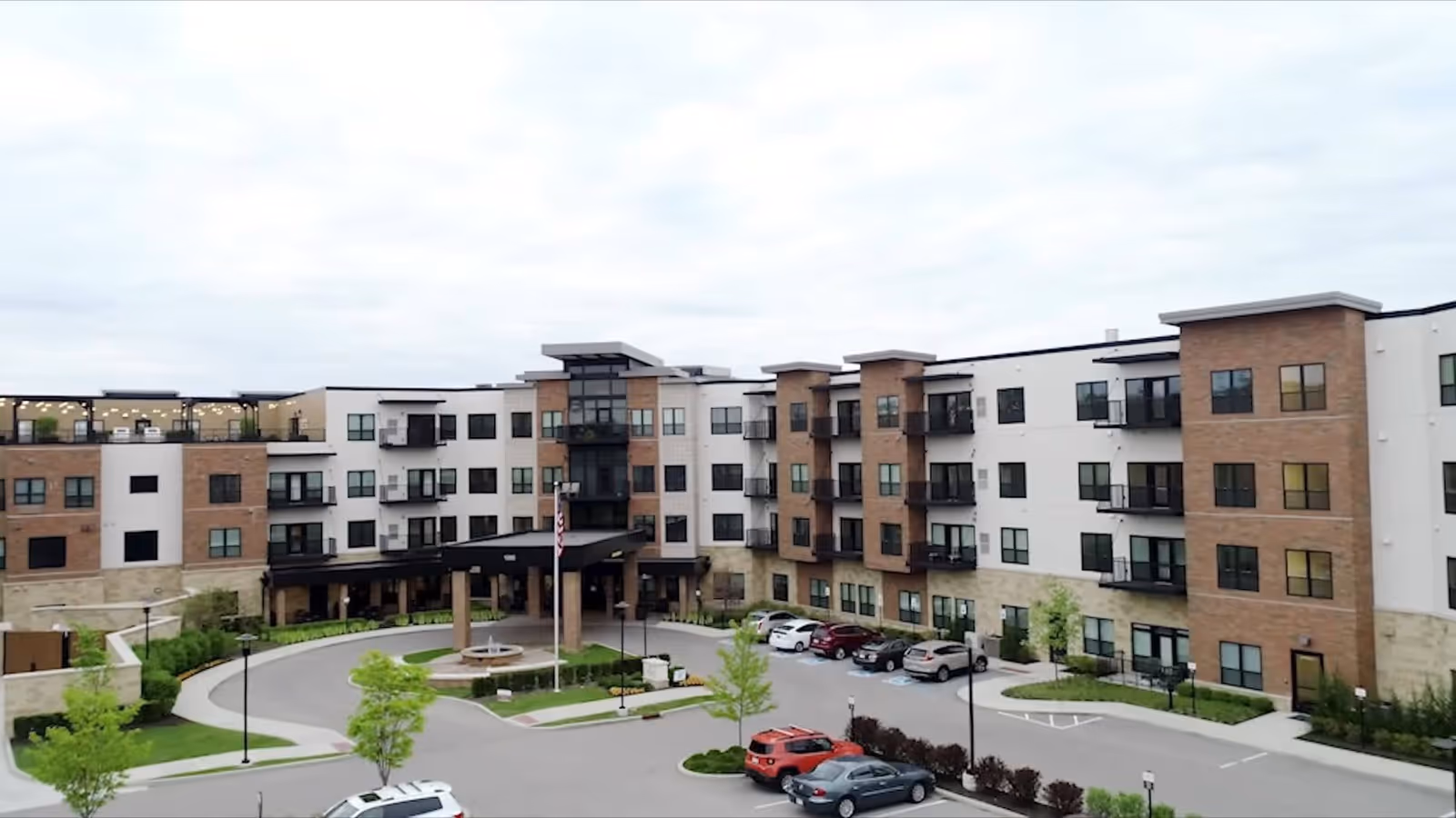 Exterior view of a modern four-story senior living facility with a circular driveway, landscaped greenery, and several parked cars under an overcast sky.