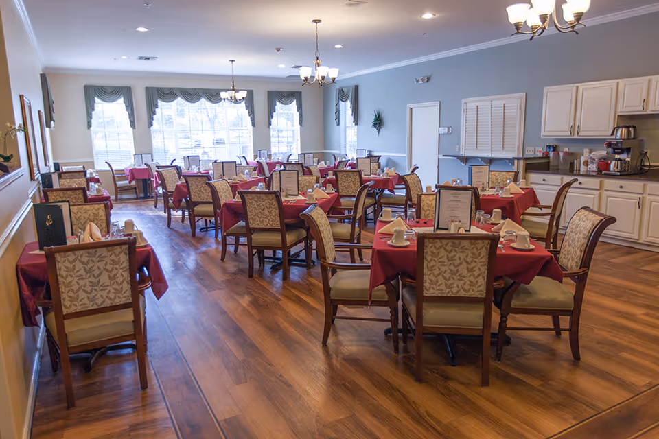 A dining room with multiple tables covered in red tablecloths, each set with menus, napkins, cups, and glasses. The room has wooden flooring, large windows with green valances, and white walls. There are chandeliers hanging from the ceiling and a counter with cabinets on the right side.