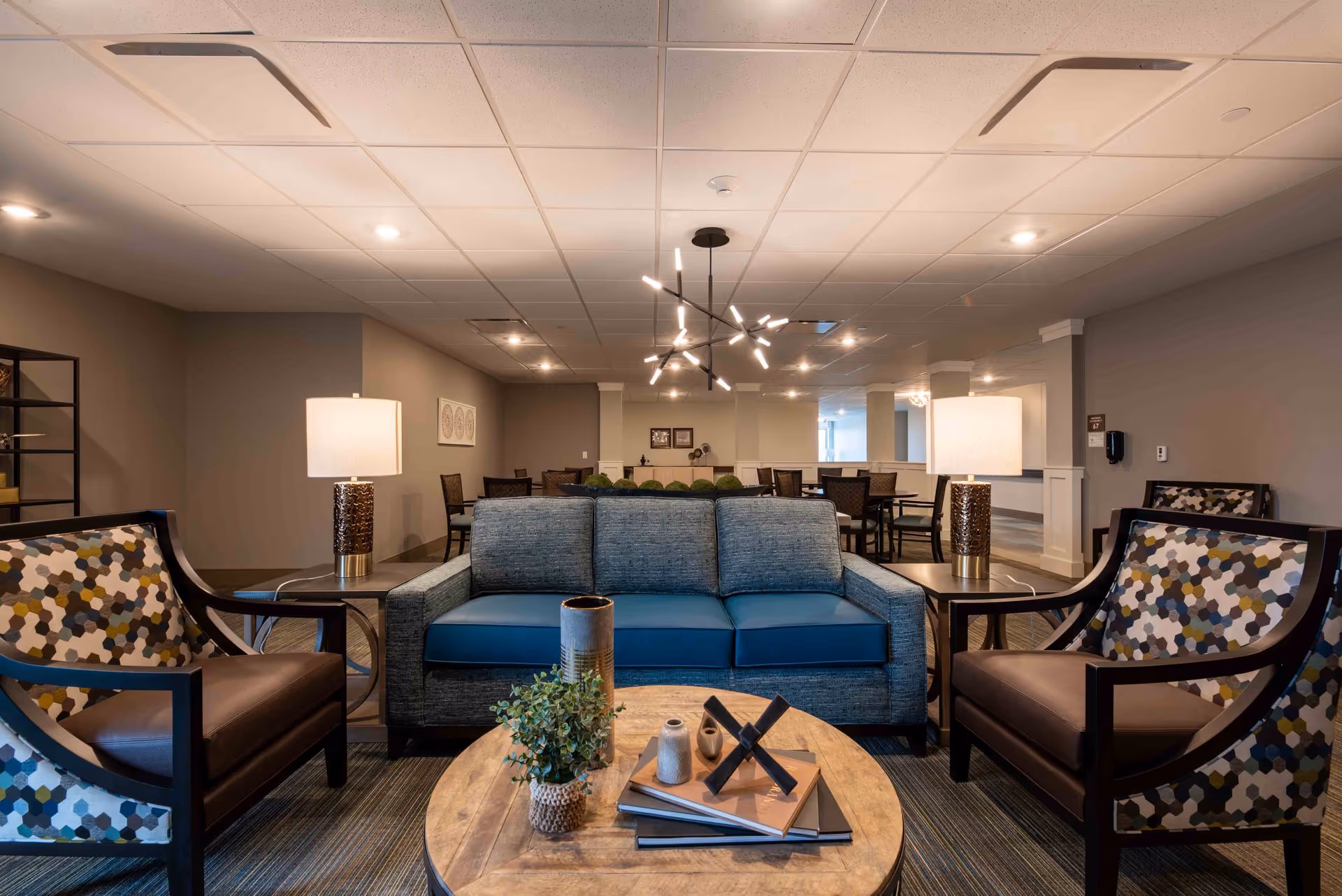 A modern senior living facility common area with a blue sofa and two patterned armchairs around a round wooden coffee table. The table has decorative items including books, a small plant, and a sculpture. Two table lamps with white shades are placed on side tables next to the sofa. In the background, there are multiple dining tables and chairs under ceiling lights, with neutral-colored walls and carpeted flooring.