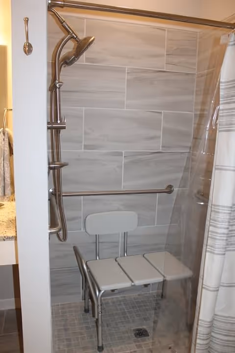 A shower area with gray tiled walls and floor, equipped with a handheld showerhead on a sliding bar, a horizontal grab bar, and a white shower chair with a backrest. A clear shower curtain with gray stripes is partially visible on the right side.