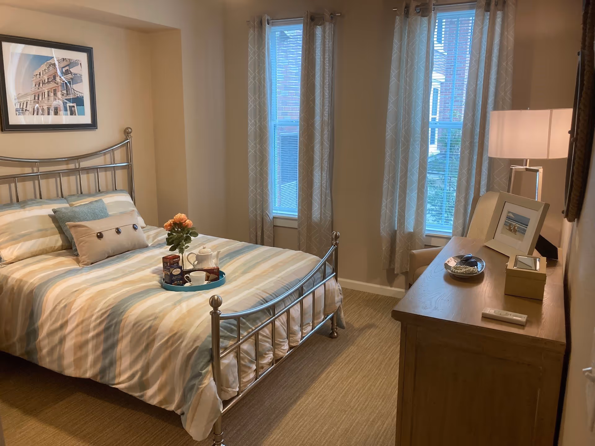 A cozy bedroom with a metal frame bed dressed in striped bedding and decorative pillows. A tray with a teapot, cup, and flowers is placed on the bed. The room has two tall windows with patterned curtains, a wooden dresser with a lamp, framed photo, and small decorative items. A framed picture hangs on the wall above the bed.
