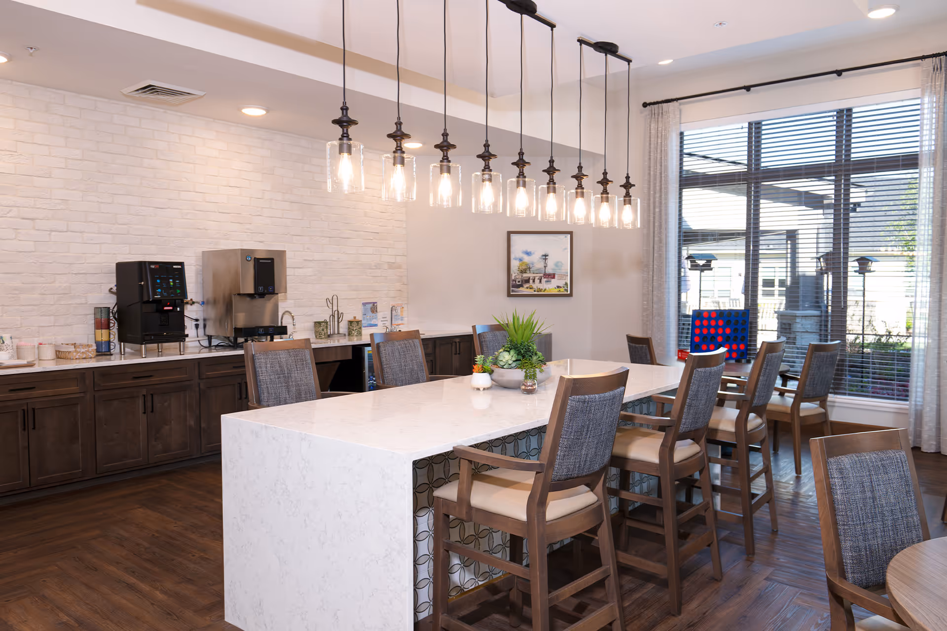 A modern dining area in a senior living facility featuring a long white marble island table with eight wooden chairs. Above the table, there are multiple hanging pendant lights with exposed bulbs. In the background, there is a countertop with coffee machines and various supplies against a white brick wall. Large windows with blinds allow natural light to enter the room, and a framed picture hangs on the wall.