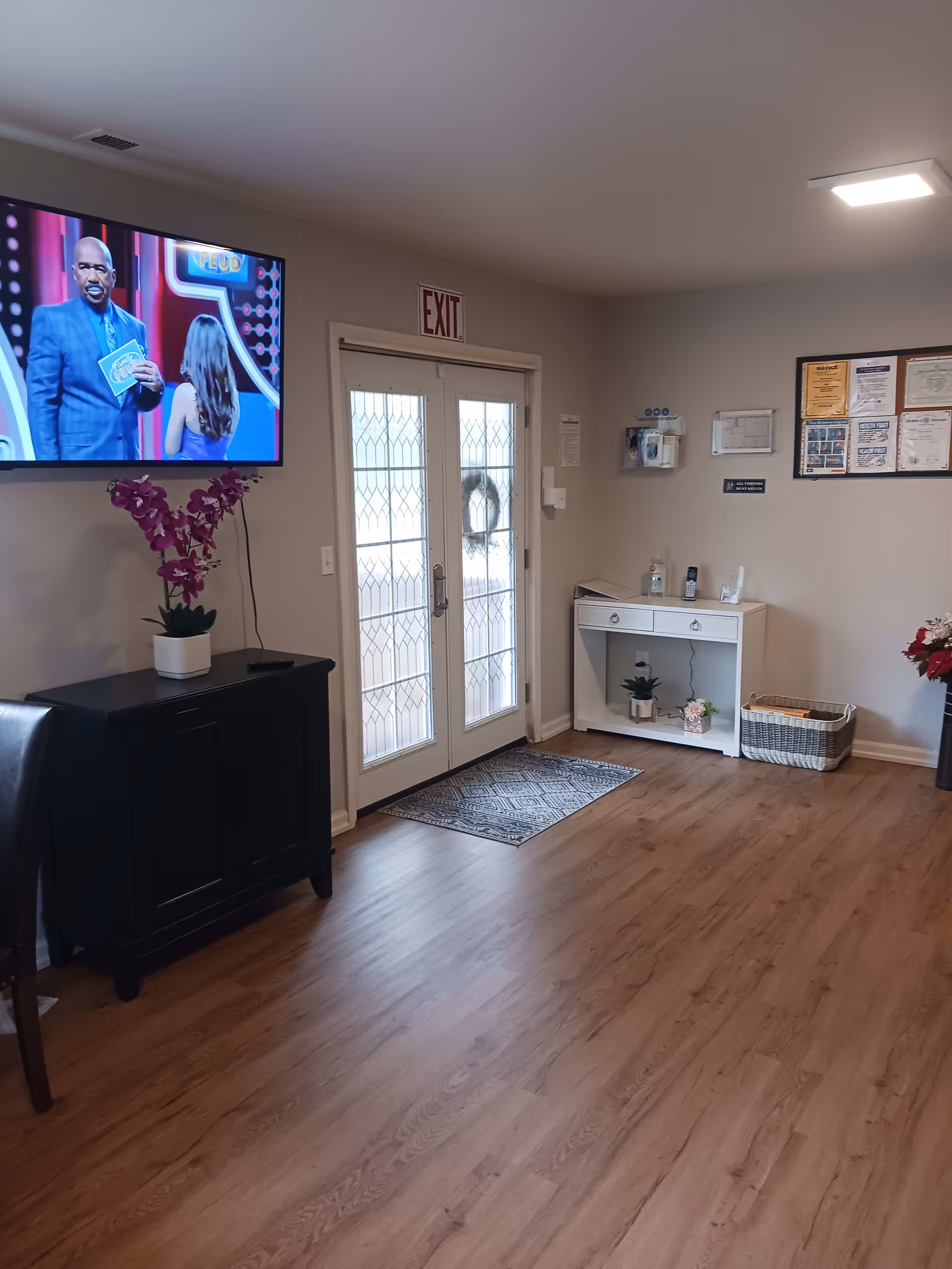 Interior view of a senior living facility common area with wooden flooring, a wall-mounted TV showing a game show, a black cabinet with a purple orchid plant, double glass doors with an exit sign above, a white console table with plants and sanitizer, and a bulletin board with notices on the wall.
