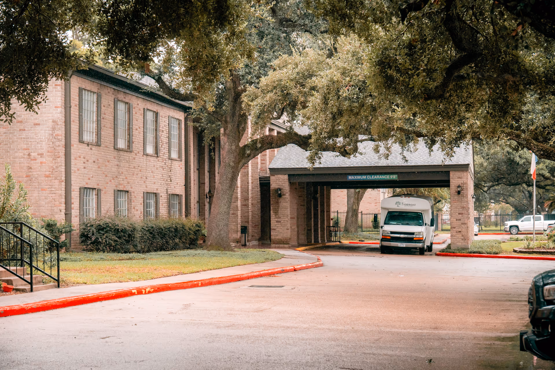 Exterior view of a brick building with multiple windows, surrounded by large trees and greenery. A covered driveway with a sign indicating maximum clearance of 9 feet 8 inches is visible, with a white van parked underneath displaying the Treemont logo. The scene includes a red-painted curb and a parking area with additional vehicles in the background.