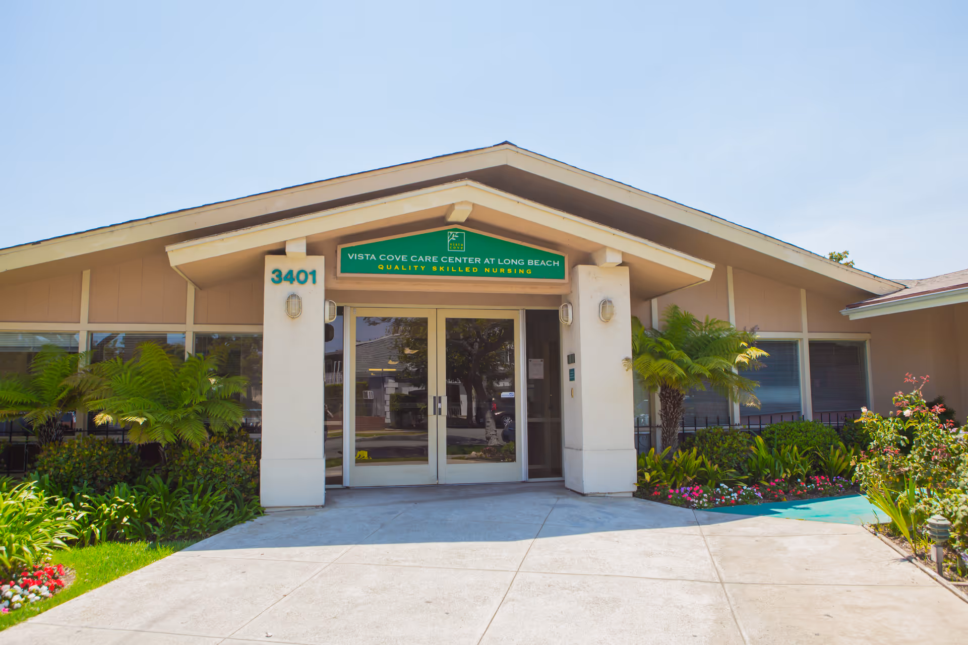 Front exterior view of a single-story healthcare facility building with a peaked roof, glass double doors entrance, and the address number 3401 displayed on a pillar. The building is surrounded by well-maintained landscaping including green plants, ferns, and colorful flowers under a clear blue sky.