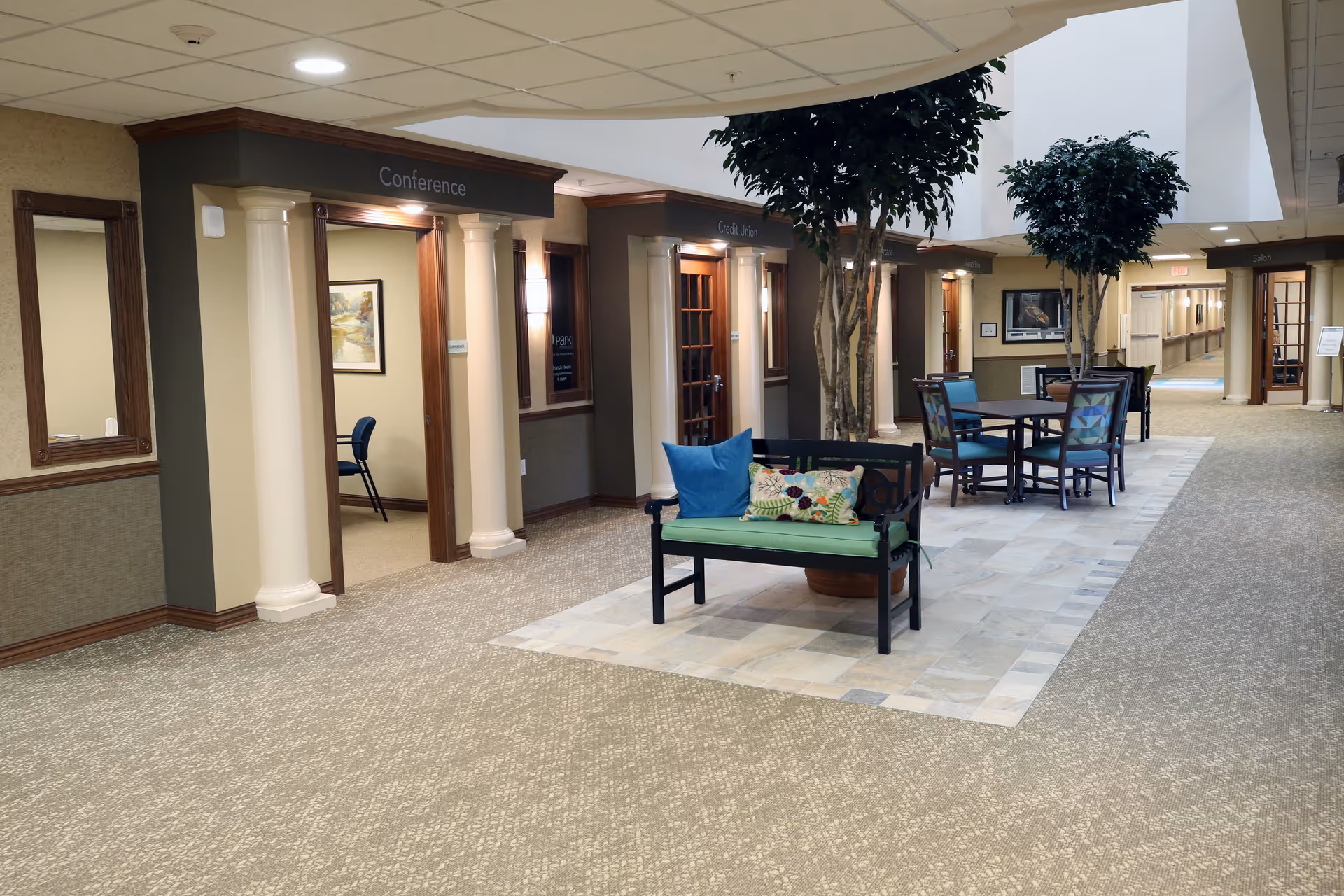 Interior hallway of a senior living facility with carpeted floors and a tiled seating area in the center featuring a bench with colorful cushions and a table with chairs. The hallway has several doorways labeled Conference, Credit Union, and Salon. There are large potted trees and framed artwork on the walls.