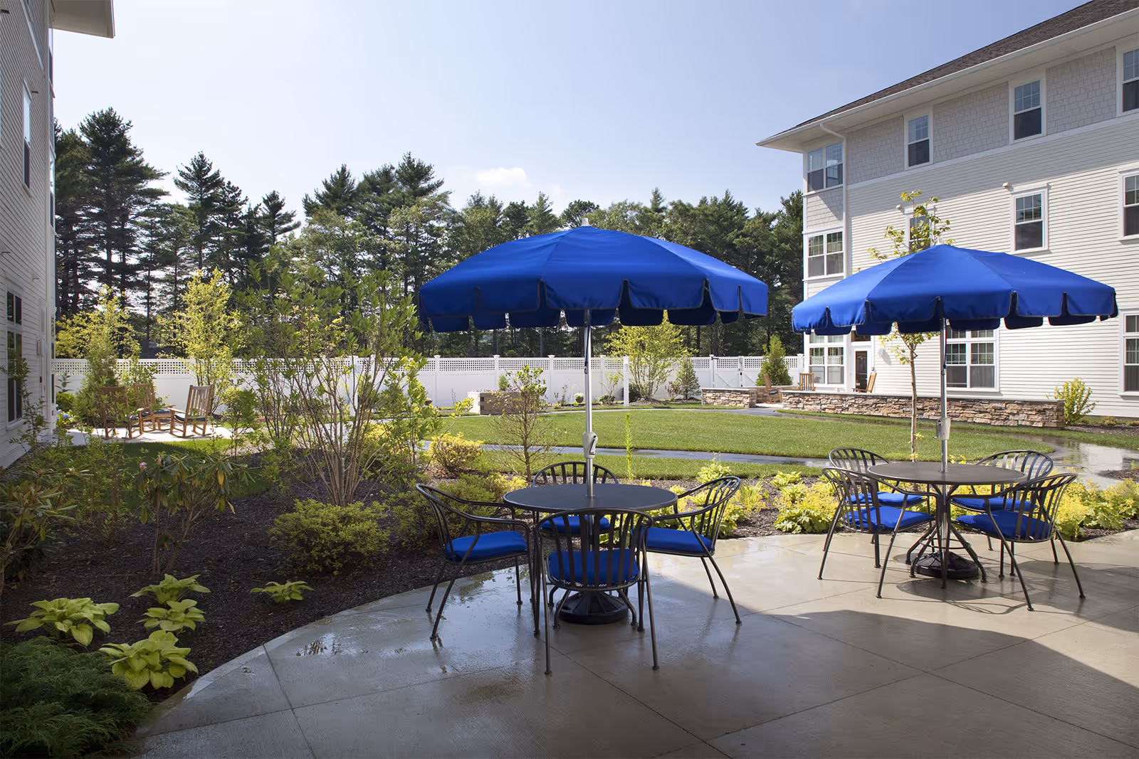 Outdoor patio area at Village At Proprietors Green with round tables and blue umbrellas, surrounded by greenery and a white fence, adjacent to a multi-story building under a clear sky.