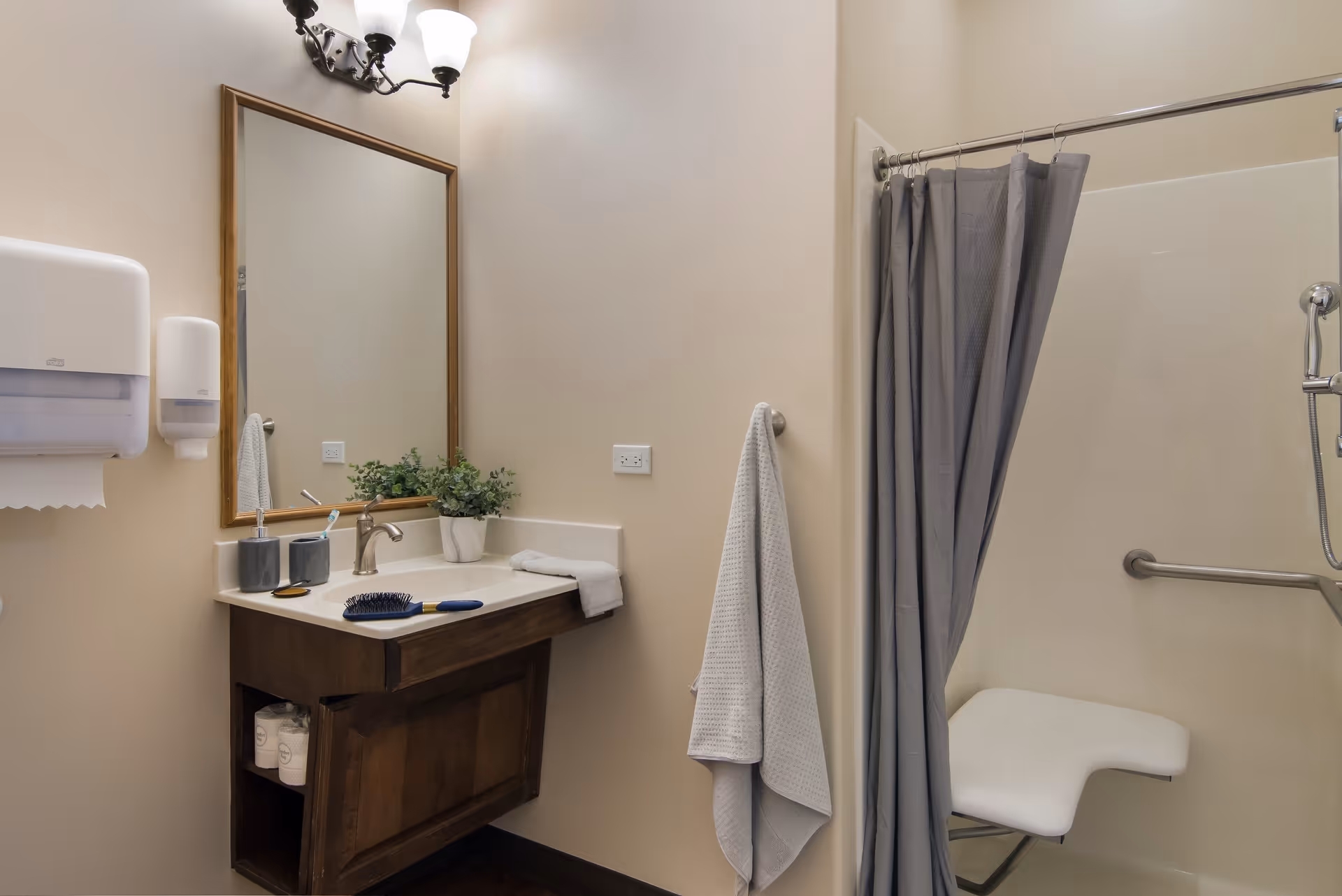 A bathroom with a wooden vanity topped with a sink, a mirror above it, a wall-mounted paper towel dispenser and soap dispenser, a towel hanging on a hook, and a shower area with a gray curtain and a white shower seat.