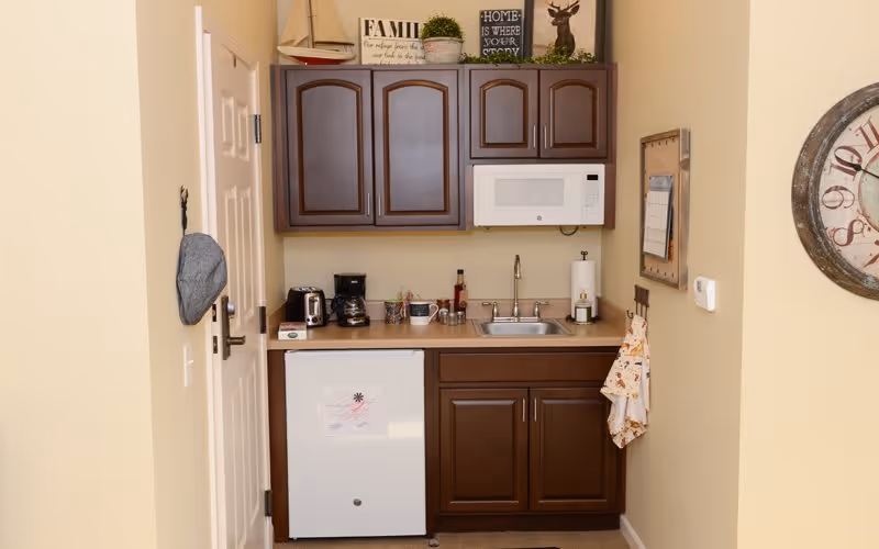 Small kitchenette with dark wood cabinets, a white microwave mounted above the sink, a mini refrigerator below the counter, a coffee maker, toaster, and various kitchen items on the countertop. Decorative signs and a small plant are placed on top of the upper cabinets. A clock is mounted on the wall to the right.