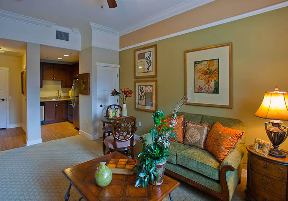 A cozy living room area in a senior living facility with a green upholstered sofa adorned with orange and patterned pillows. A wooden coffee table with decorative items and a potted plant sits in front of the sofa. To the right, there is a wooden side table with a lamp and a framed photo. The walls are decorated with framed floral artwork. In the background, a small dining area with two wooden chairs and a round table is visible, along with a kitchen area featuring dark wood cabinets and stainless steel appliances.