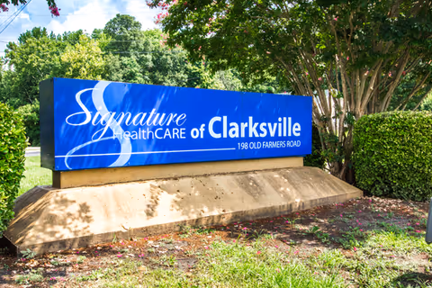 A blue and white outdoor sign for Signature HealthCARE of Clarksville located at 198 Old Farmers Road, surrounded by green bushes and trees under a partly cloudy sky.