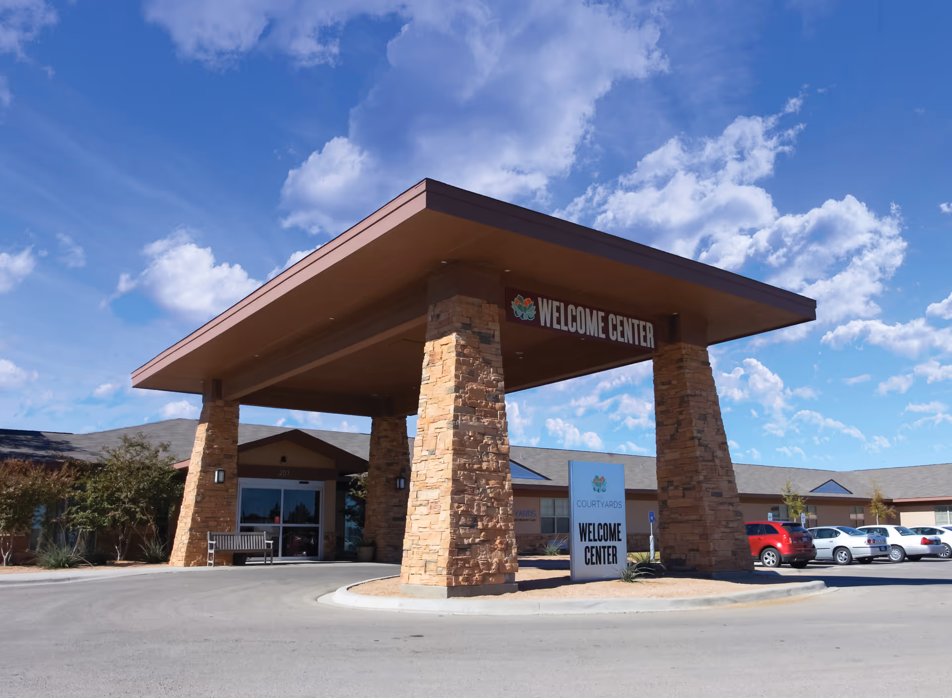 Exterior view of The Courtyards Assisted Living and Memory Care Welcome Center with a large covered entrance supported by stone pillars, a sign reading 'Welcome Center', several parked cars, and a bright blue sky with scattered clouds.