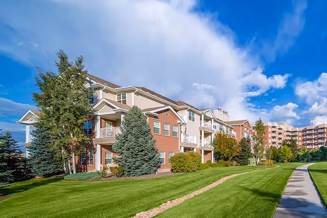 Exterior view of a senior living facility with well-maintained green lawns, trees, and a paved walkway under a partly cloudy blue sky.