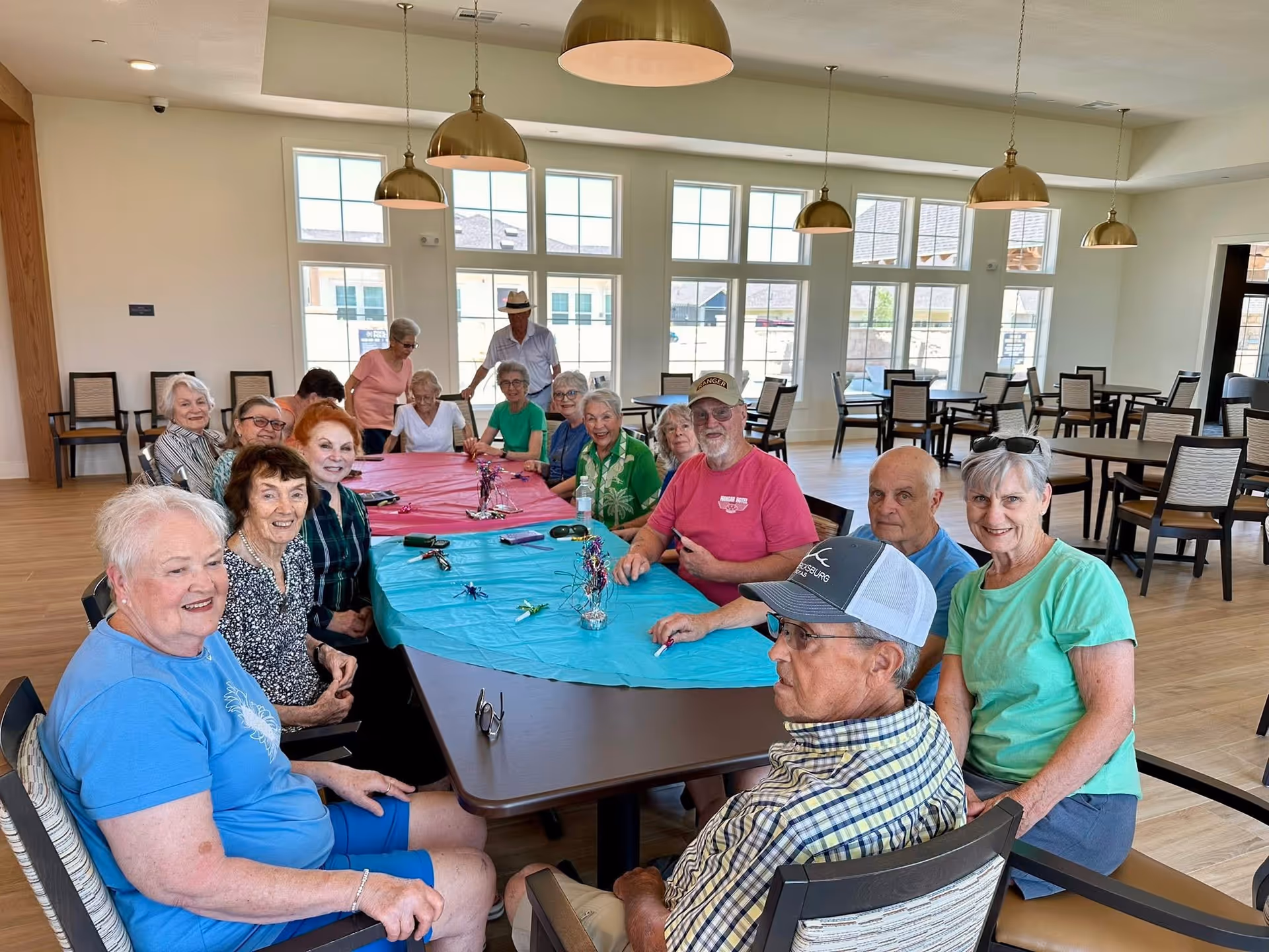 A group of elderly people sitting around a large table covered with blue and pink tablecloths in a bright, spacious room with large windows and hanging pendant lights. They appear to be socializing and enjoying each other's company.