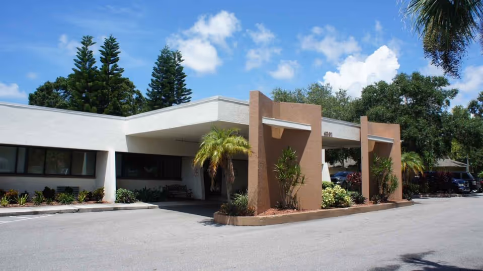 Exterior view of Bay Pointe Nursing Pavilion showing the entrance with a covered drop-off area, surrounded by landscaped plants and trees under a blue sky with some clouds.