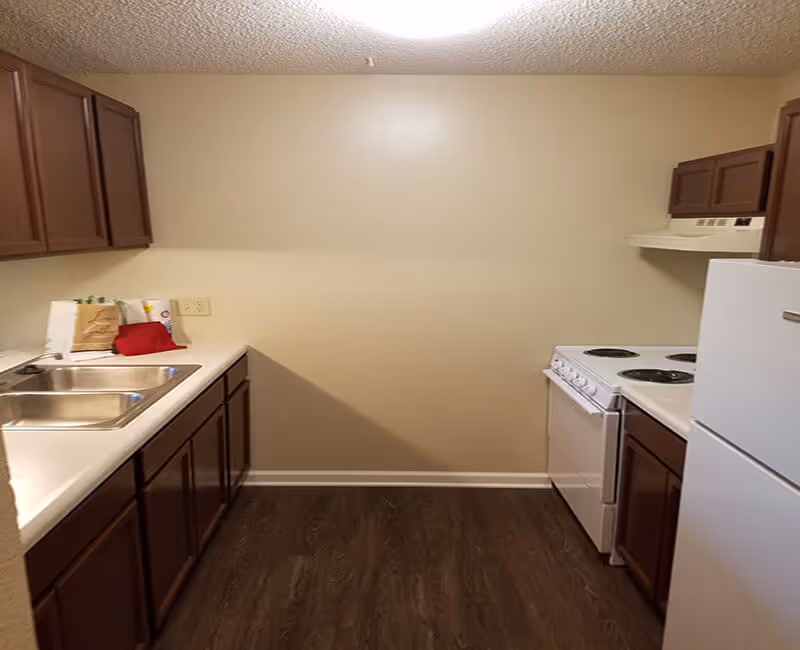 A small kitchen with dark wood cabinets, white countertops, a double sink, a white electric stove with four burners, a white refrigerator, and a beige wall. The floor is dark wood, and there are a few items on the counter including a paper bag and a red cloth.