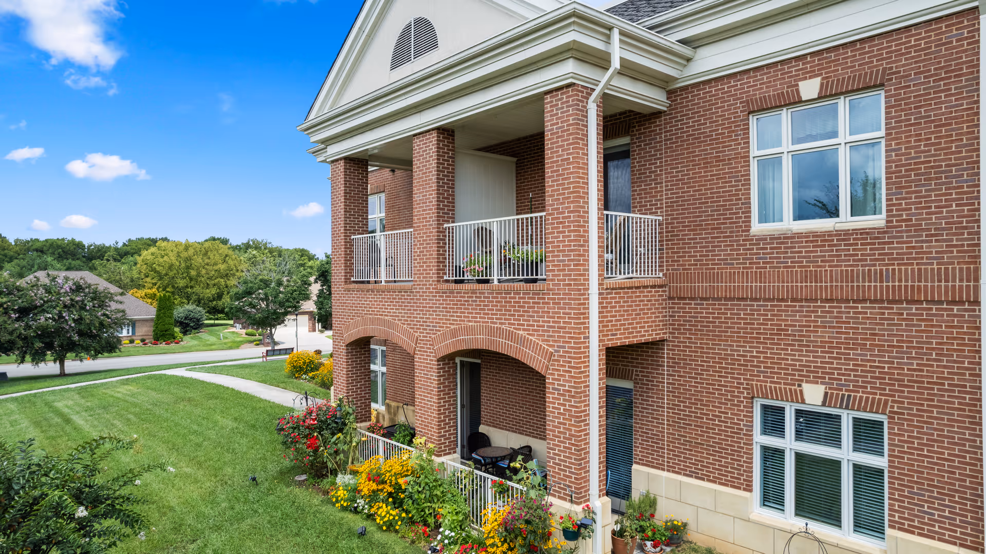 Exterior view of a two-story brick building with balconies and a well-maintained lawn with colorful flowers and shrubs under a blue sky with a few clouds.