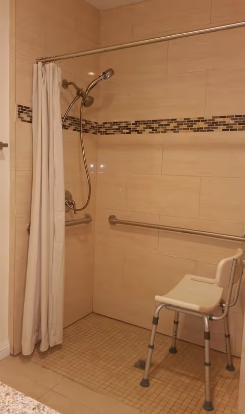 Tiled roll-in shower with a handheld showerhead, grab bars, shower curtain, and a shower chair.