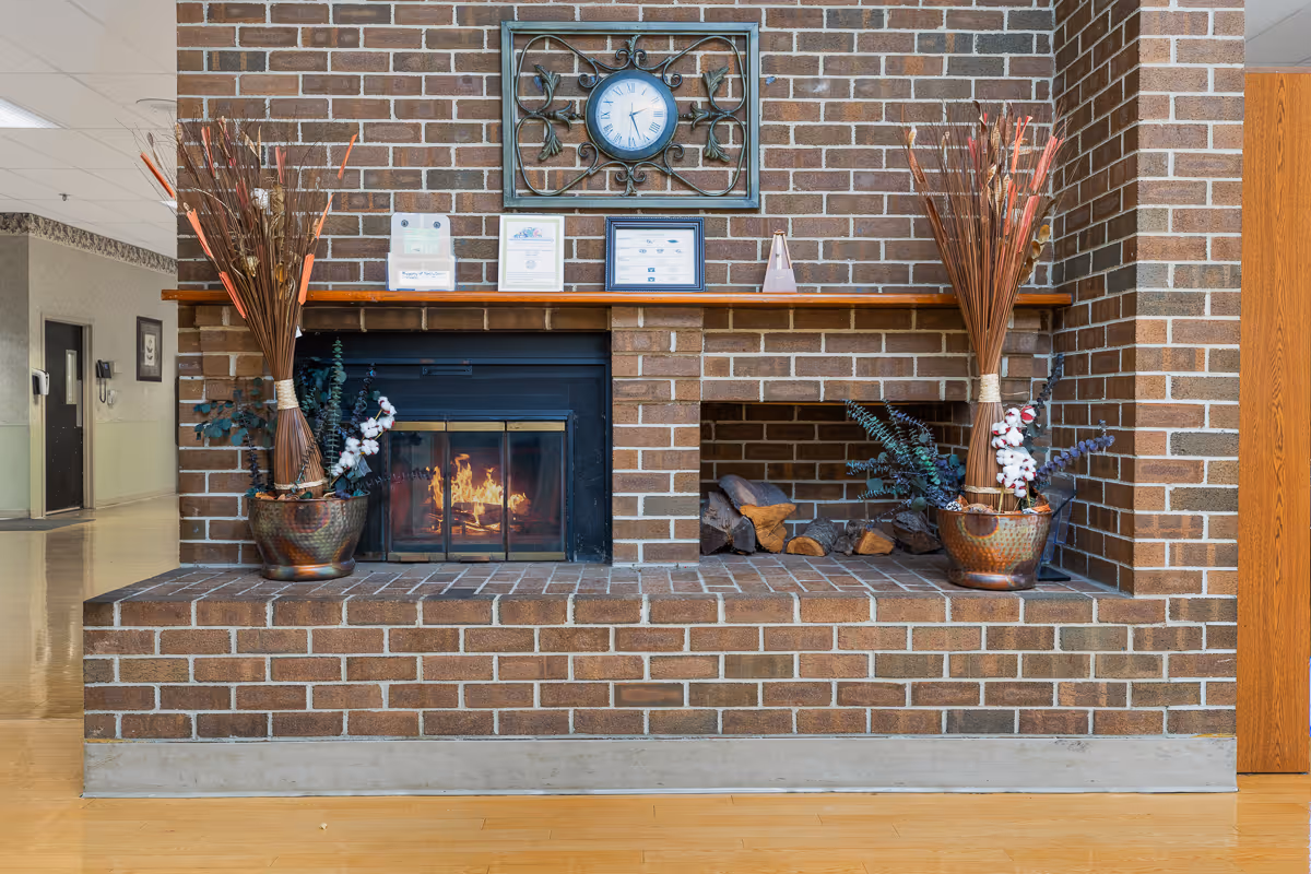 A brick fireplace with a fire burning inside on the left side and stacked firewood on the right side. Above the fireplace is a wooden mantel with framed certificates and a decorative clock mounted on the brick wall. Two large vases with tall dried floral arrangements are placed on either side of the fireplace. The floor is wooden, and a hallway is visible to the left.
