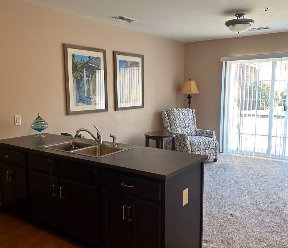 Open-plan living area with a kitchen island and sink in the foreground, an upholstered armchair and side table near sliding glass doors.
