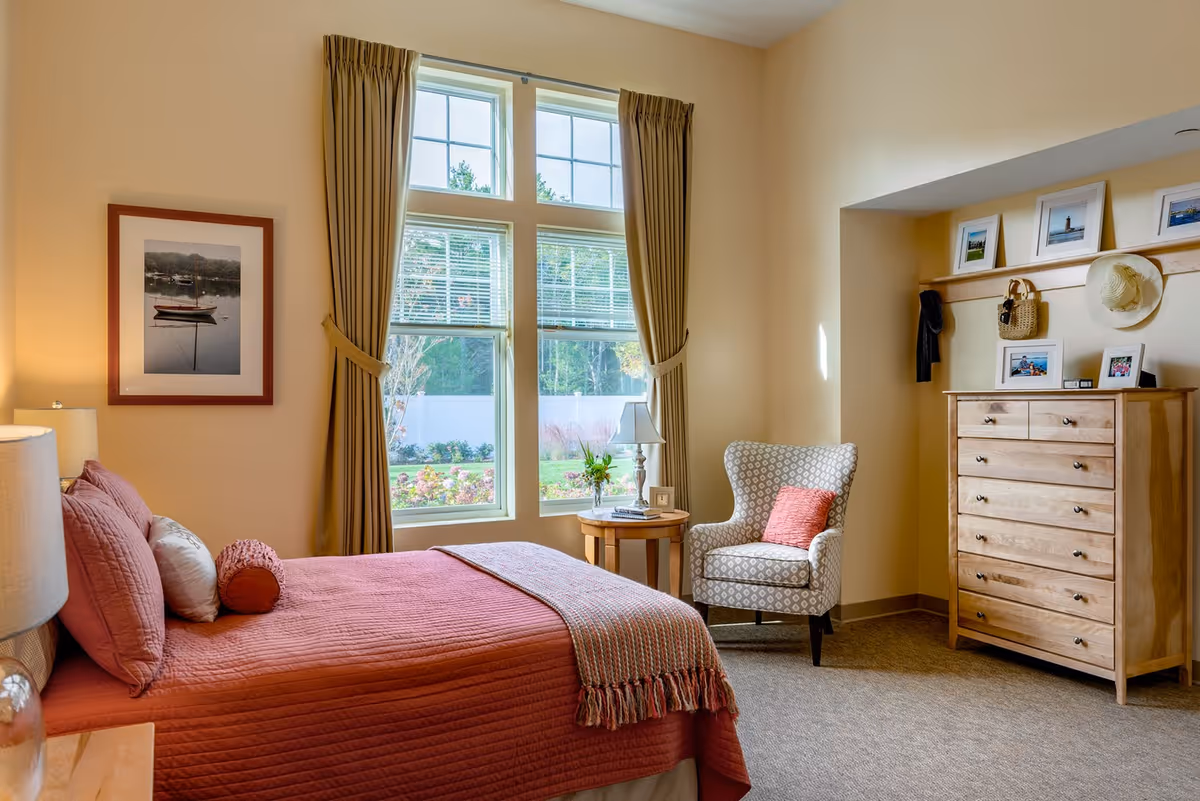 A cozy bedroom with a bed covered in a rust-colored quilt and pillows, a wooden nightstand with a lamp, a framed picture of a boat on the wall, a window with beige curtains letting in natural light, a round wooden side table with a lamp and a small plant, a patterned armchair with a coral cushion, and a wooden dresser with framed photos and a hat hanging above it.