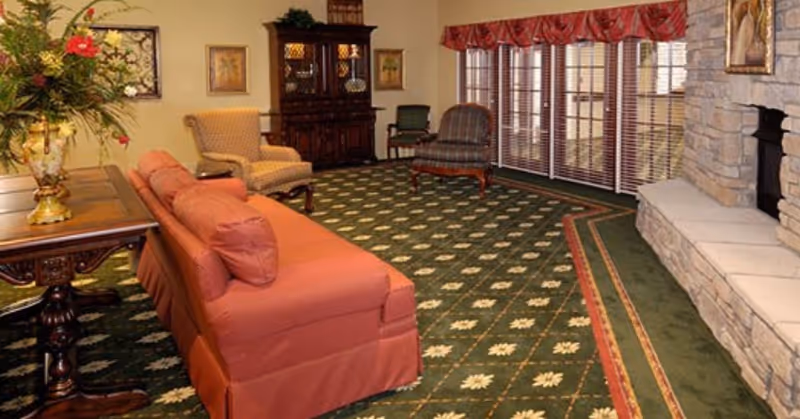 A cozy living room with a patterned green carpet, a red sofa, two armchairs, a wooden table with a floral arrangement, a wooden cabinet, and a stone fireplace. Large windows with blinds and red valances allow natural light into the room.