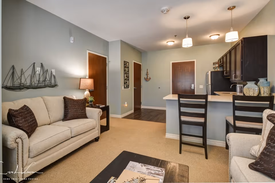 Interior view of a senior living apartment featuring a beige sofa with dark brown pillows, a coffee table with magazines, a kitchen area with a breakfast bar and two chairs, dark wood cabinets, and a refrigerator. The walls are painted light gray with decorative wall art and a lamp on a side table next to the sofa. There are three wooden doors visible, and the flooring transitions from carpet in the living area to wood in the kitchen and hallway.