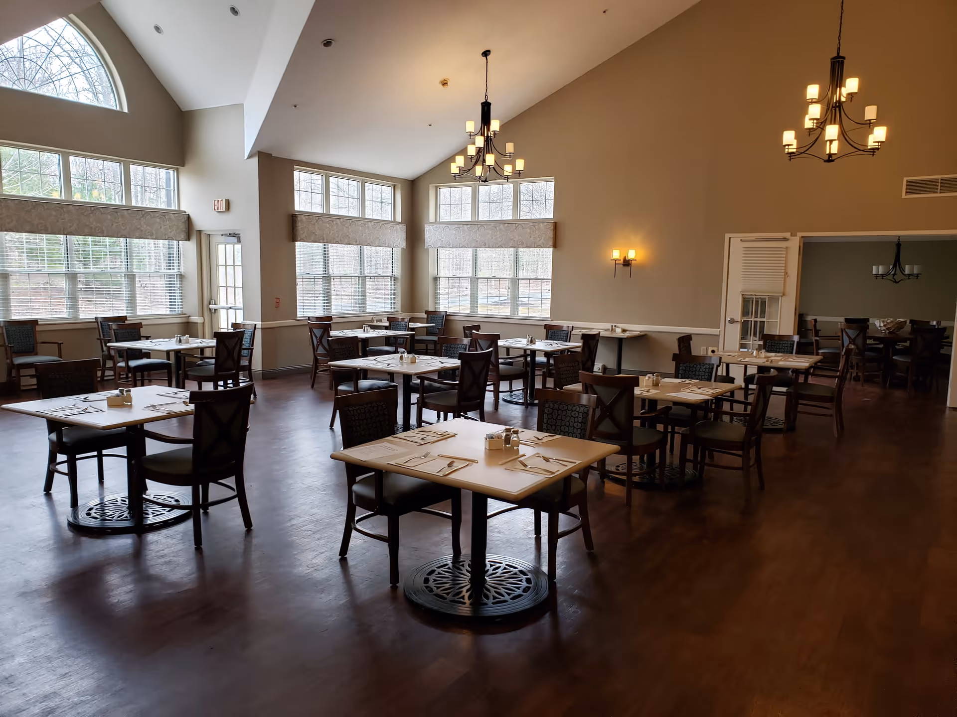 A spacious dining room with multiple tables and chairs arranged neatly. Each table is set with placemats, napkins, and utensils. Large windows with blinds allow natural light to fill the room. Two chandeliers hang from the ceiling, and there are wall-mounted lights on the beige walls. The floor is wooden, and there is a door leading to another room in the background.