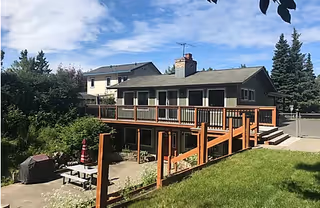 Exterior view of a two-story residential building with a large wooden deck and railing, surrounded by green grass, trees, and a clear blue sky with some clouds.