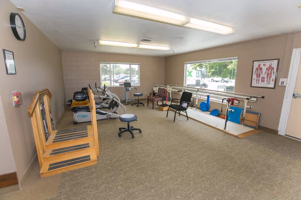 A rehabilitation room with exercise equipment including stationary bikes, parallel bars for walking practice, chairs, and a wooden step platform. The room has beige walls, two large windows showing a parking lot outside, a clock on the wall, and a poster of the muscular system.