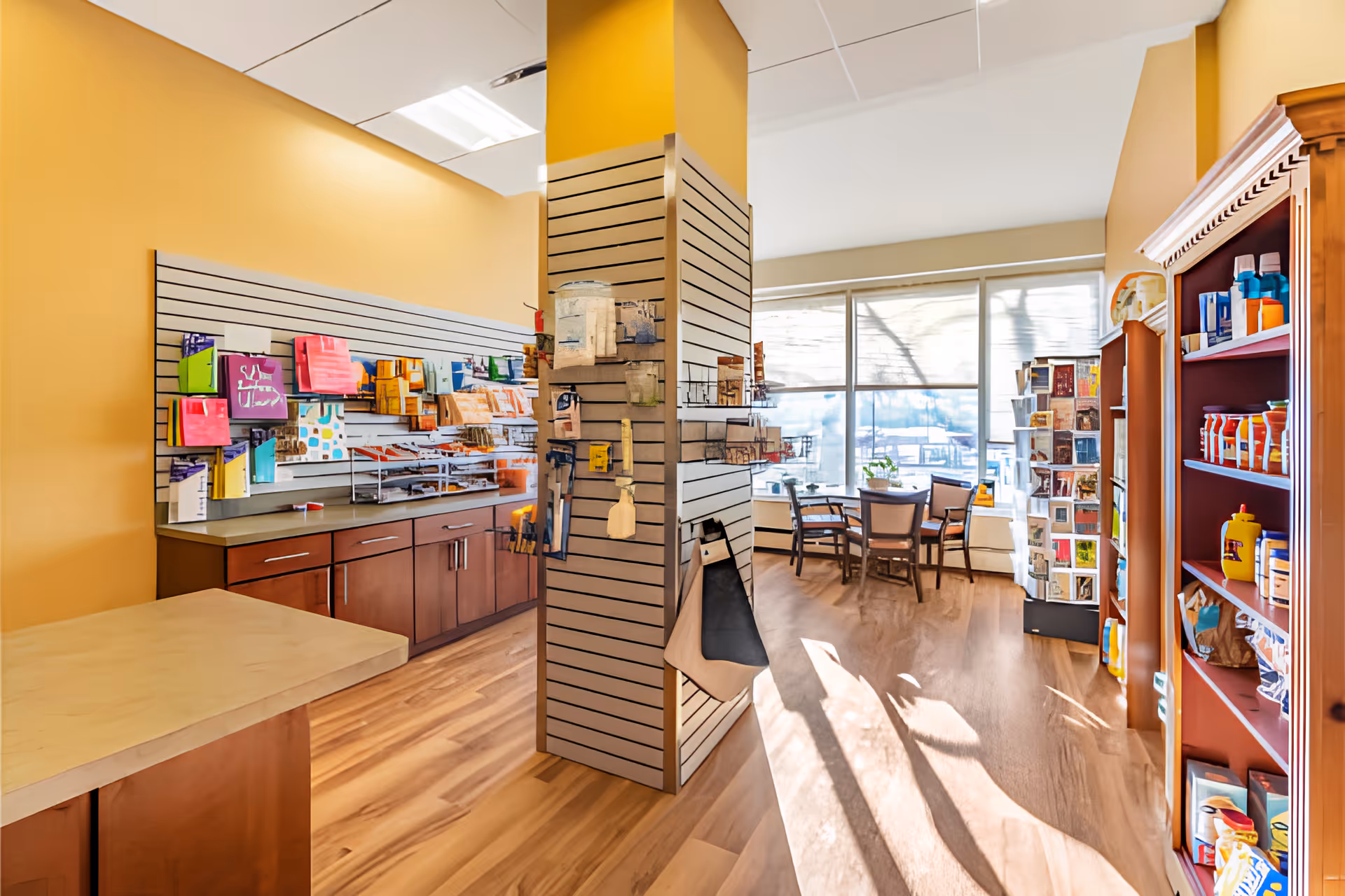Interior view of a small shop or convenience area within a senior living facility. The space features wooden shelves stocked with various items such as snacks, condiments, and greeting cards. There is a counter with cabinets below and a display wall with hanging merchandise. In the background, a seating area with a table and chairs is visible near large windows letting in natural light.