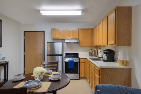 Small open kitchen with wooden cabinets, stainless refrigerator and stove, and a round dining table set for four.