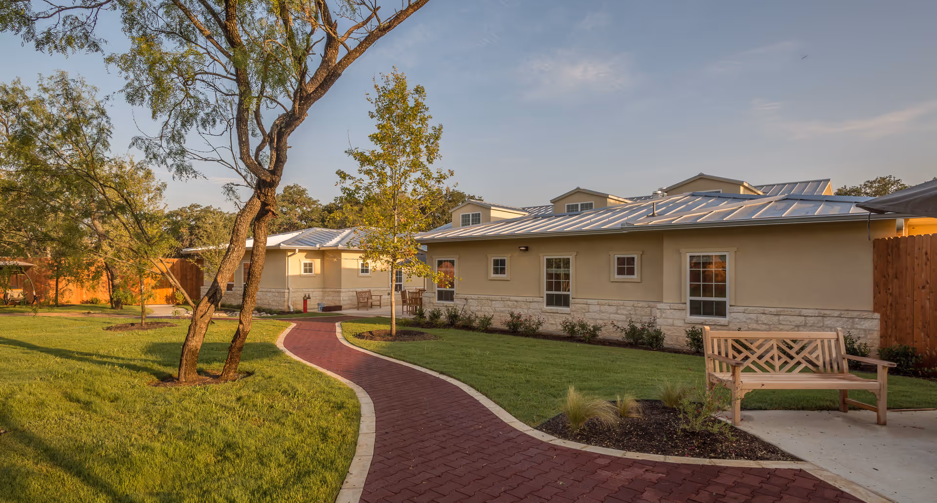 A landscaped outdoor area at Seasons Memory Care at Shavano Park featuring a winding red brick pathway, green grass, trees, and a wooden bench near a beige building with multiple windows and a metal roof under a partly cloudy sky.