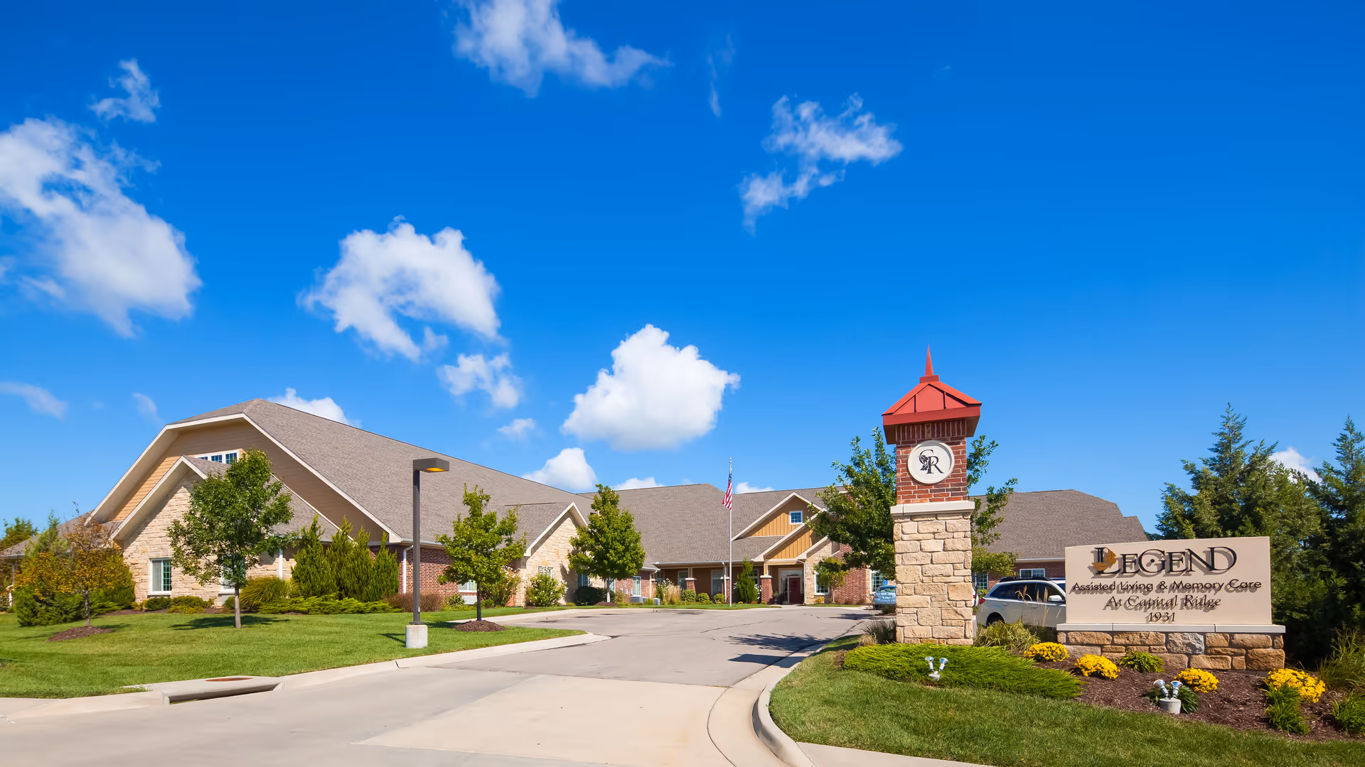Exterior view of Legend at Capital Ridge, a senior living facility with a large building, well-maintained landscaping, trees, and a clear blue sky with some clouds. The entrance sign reads 'Legend Assisted Living & Memory Care At Capital Ridge 1991'.