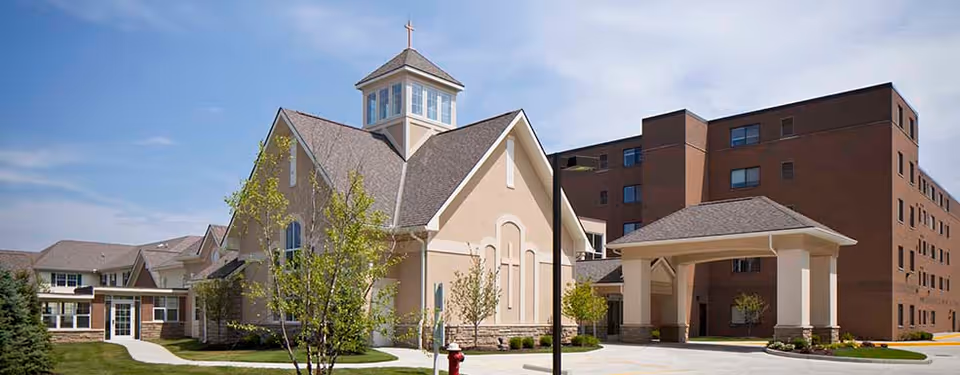 Exterior view of Ohio Living Swan Creek facility showing a beige chapel-like building with a cross on top, adjacent to a larger multi-story brick building. The scene includes a covered entrance, a sidewalk, green grass, and small trees under a blue sky with some clouds.