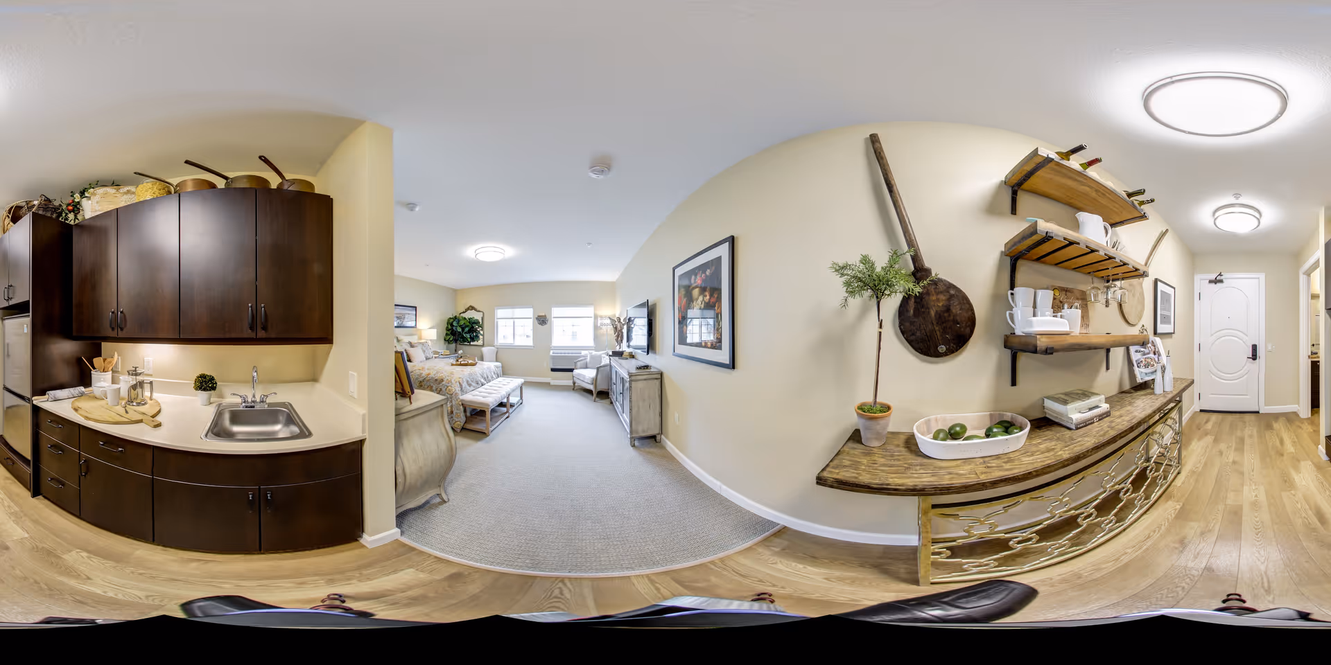 A wide-angle view of a senior living facility apartment featuring a small kitchen area with dark wood cabinets and a sink on the left, a cozy living space with a bed, armchair, and TV in the center, and a hallway with decorative shelves and a front door on the right. The room has light-colored walls and wood flooring, with soft lighting from ceiling fixtures.
