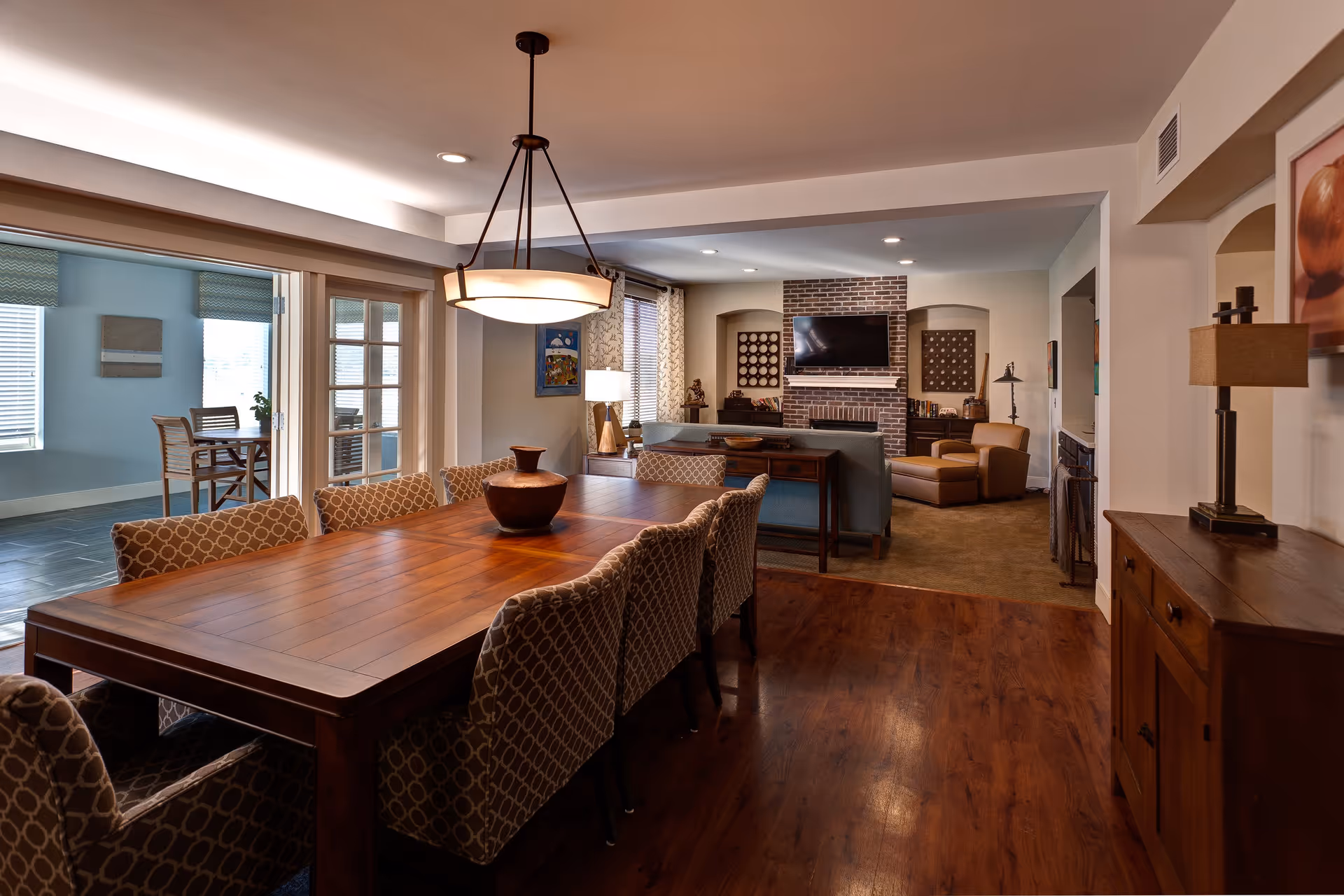 A spacious dining room featuring a large wooden table surrounded by patterned chairs, with a view into a cozy living area that includes a brick fireplace and a television.