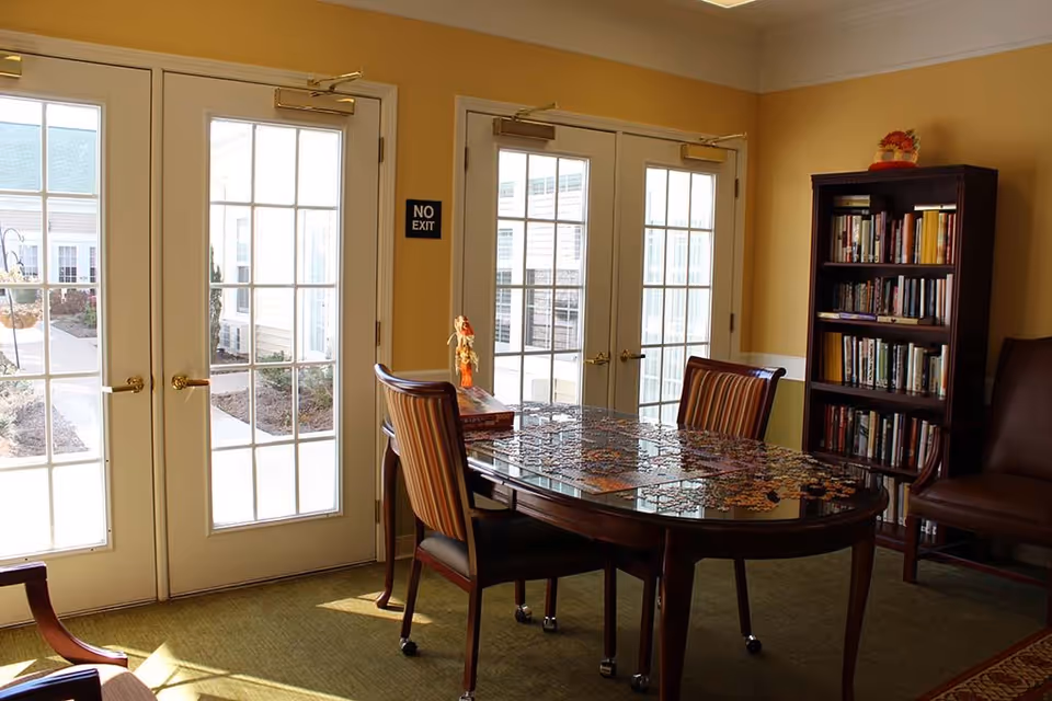 A cozy room with a wooden table in the center covered with a partially completed jigsaw puzzle. Two striped chairs are positioned around the table. Behind the table is a tall bookshelf filled with books, and to the right is a brown leather chair. The room has large glass double doors letting in natural light, with a 'NO EXIT' sign on the wall between the doors. The walls are painted yellow with white trim, and the floor is carpeted in green.