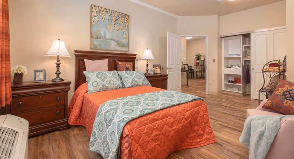 A cozy bedroom with a wooden bed frame, an orange quilted bedspread, and blue patterned pillows and throw. There are two wooden nightstands with lamps and decorative items on either side of the bed. A painting hangs above the bed. The room has wood flooring, an open closet with shelves and hanging space, and a doorway leading to another room with a desk and chair.