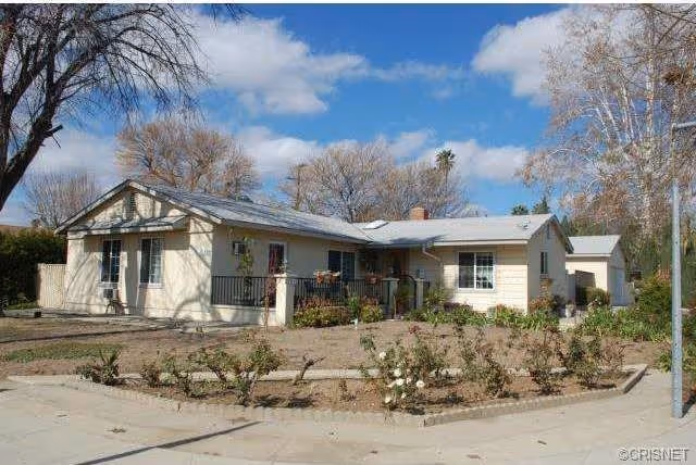 Single-story residential building with light-colored exterior walls and a gray roof, surrounded by a garden with small bushes and trees under a partly cloudy blue sky.